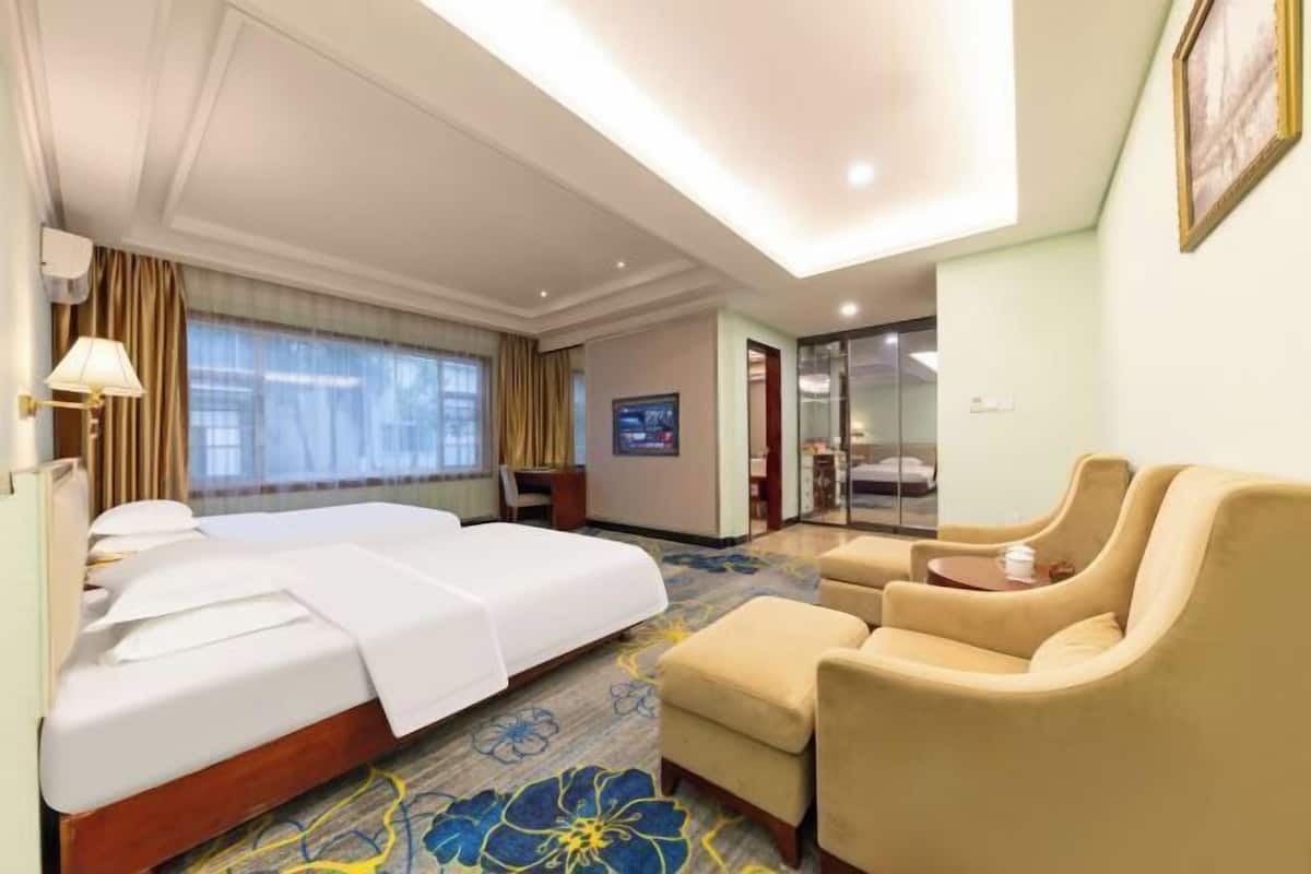 A hotel room with a bed, two armchairs, a small table, a desk, and a large window with curtains.
