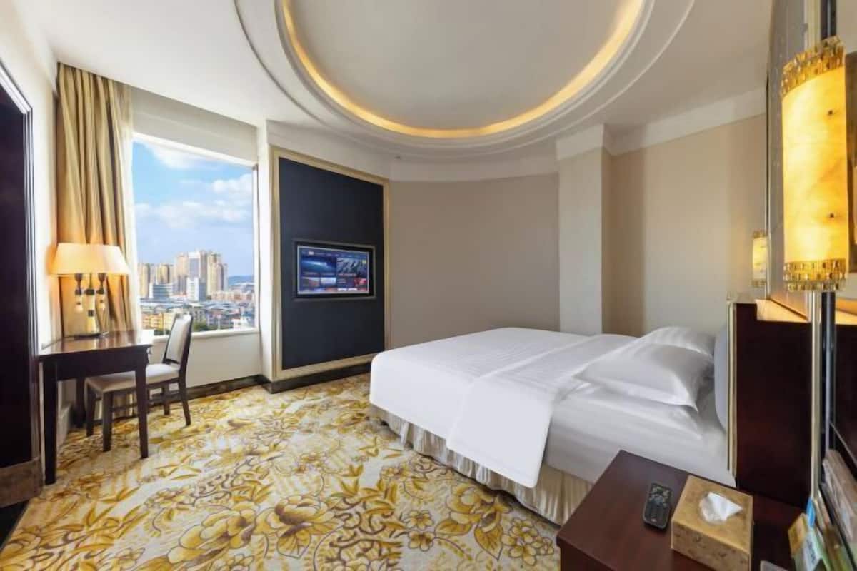 A hotel room with a bed, a desk, a chair, a TV, and a large window with a city view.