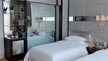 A hotel room with two beds, a mirrored wardrobe, a bedside table with a lamp, and a small shelf with decorative items.