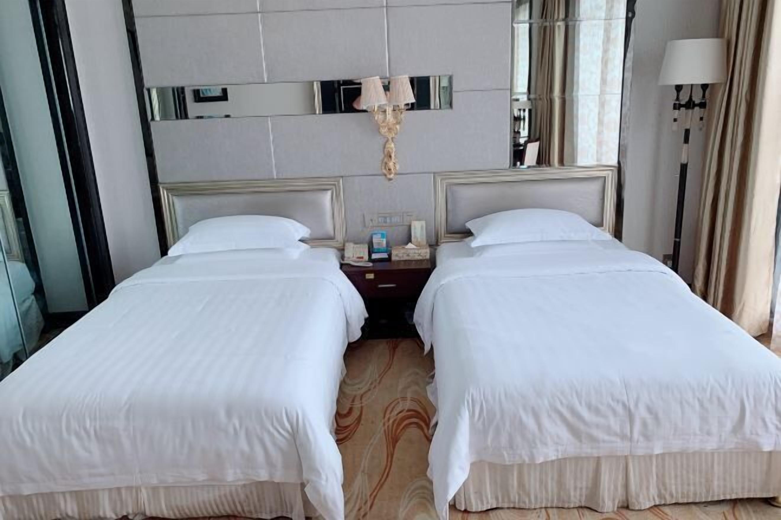 A hotel room with two single beds, a large mirror above the beds, a nightstand with a lamp, and a window with curtains.