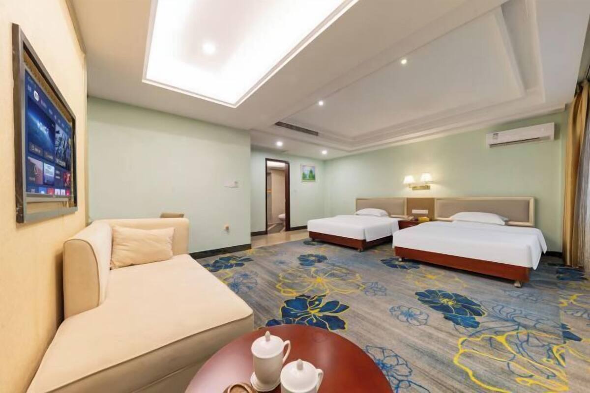 A hotel room with two beds, a sofa, a small table with a teapot, and a ceiling light.