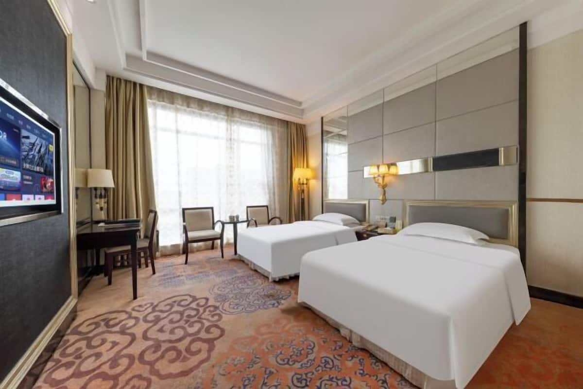 A hotel room with two beds, a desk with a chair, a TV, and a large window with curtains.