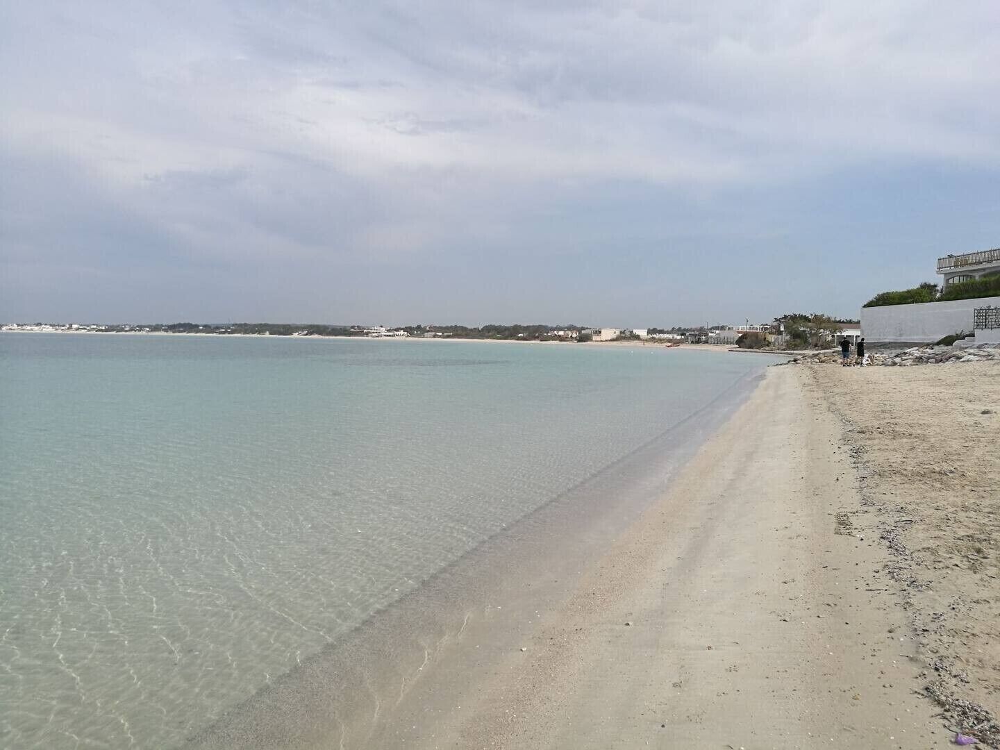 Una playa con agua cristalina, una orilla arenosa y edificios al fondo.