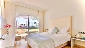 Un dormitorio con una cama grande, una mesita de noche con un reloj y una manzana, un taburete pequeño, y vistas a barcos y palmeras a través de un ventanal amplio.