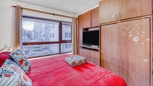 Una camera da letto con una coperta rossa, un armadio in legno e un'ampia finestra con vista sugli edifici.