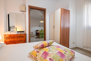 Une chambre à coucher comprenant un lit, une commode, un miroir, une armoire et donnant sur la salle à manger.