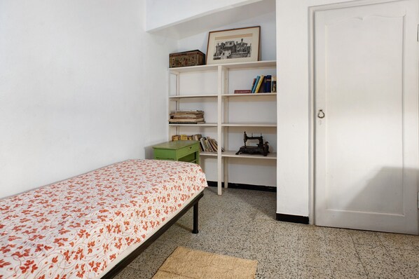 Una stanza piccola con un letto singolo, un comodino verde, una libreria bianca con libri e una cornice, e una porta.