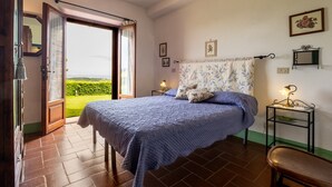 A bedroom with a bed, a nightstand, a chair, a window with curtains, and a view of greenery outside. - Apartment "Lavanda" with Mountain View, Shared Pool & Wi-Fi (radicofani)