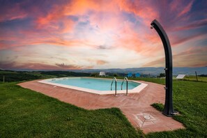 A swimming pool with a curved structure, surrounded by a paved area and grass, with a scenic view of hills and a sunset sky. - Apartment "Lavanda" with Mountain View, Shared Pool & Wi-Fi (radicofani)