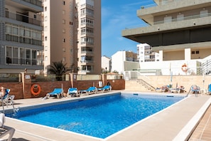 A swimming pool with lounge chairs, a brick wall, and high-rise buildings in the background. - Beach Front Holiday Studio Apartment with Sea Views, Private Terrace, and Wi-Fi (Fuengirola)