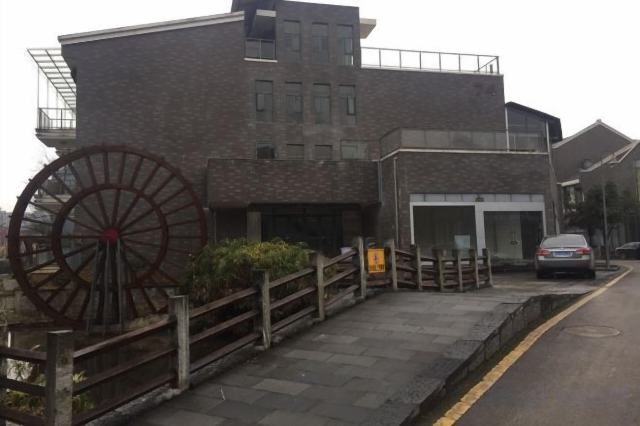 A building with a large water wheel, a paved entrance, and a car parked on the street.