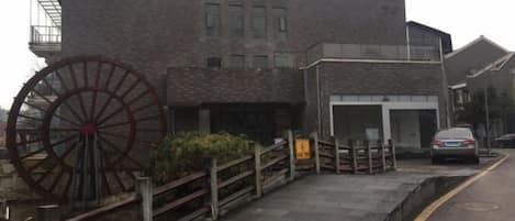A building with a large water wheel, a paved entrance, and a car parked on the street.