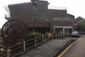 A building with a large water wheel, a paved entrance, and a car parked on the street. - Lishuiwan Hotel (Luoping)