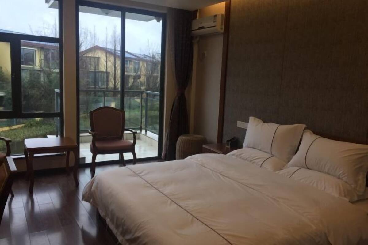 A hotel room with a large bed, a chair, a small table, and a view through a glass door to the outside.