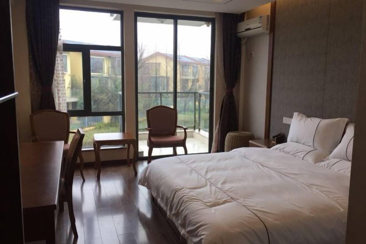 A hotel room with a large bed, a desk, a chair, and a view of a residential area through the window.
