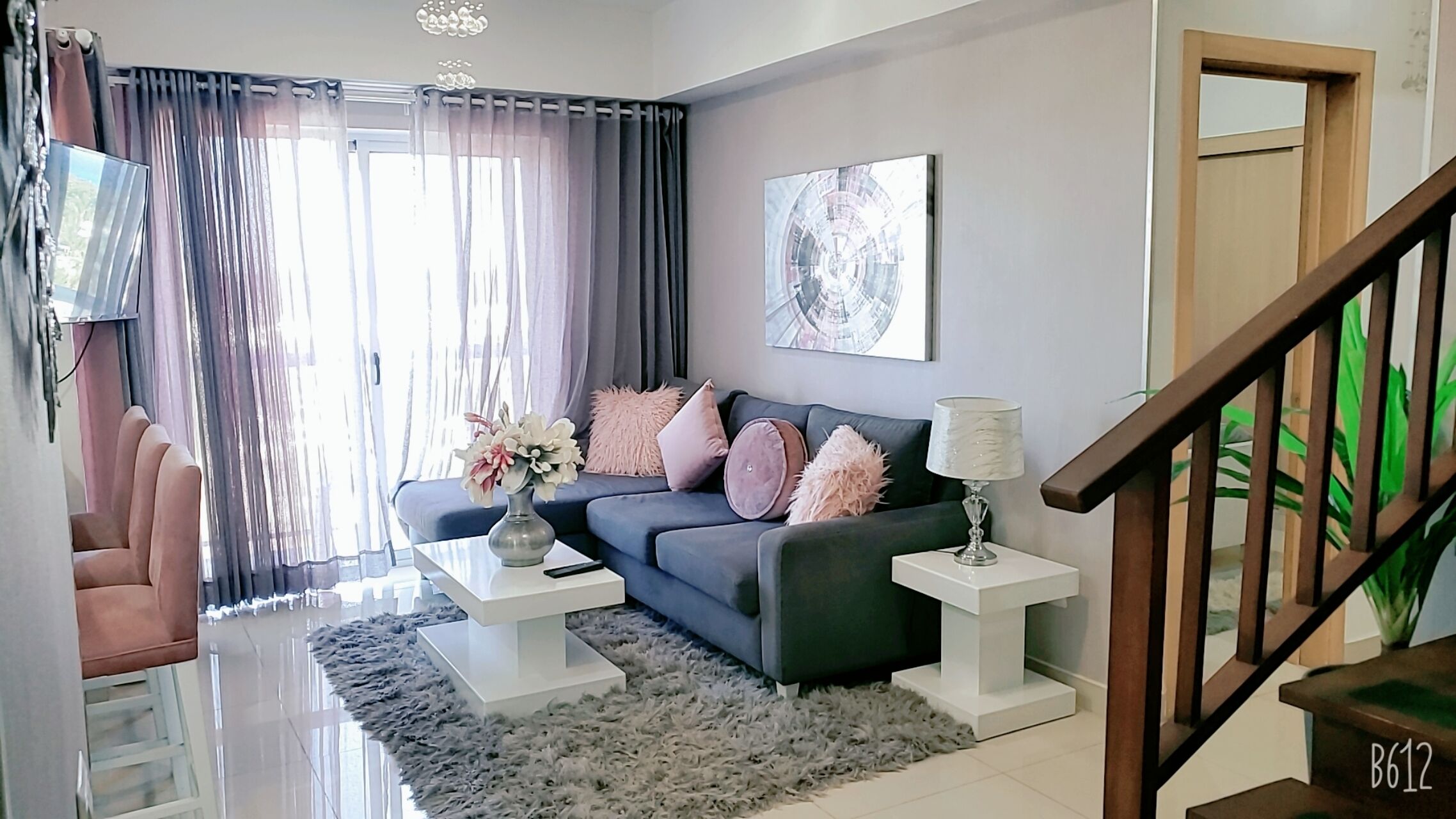 A modern living room with a dark blue sofa, white side tables, fluffy grey rug, and a staircase with wooden railings.