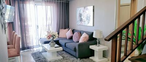 A modern living room with a dark blue sofa, white side tables, fluffy grey rug, and a staircase with wooden railings.