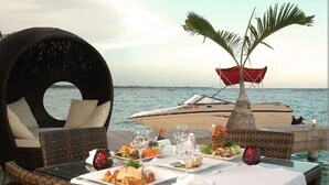 A dining table with plates of food and glasses, a wicker chair, and a boat in the background. - Hotel Boca del Mar Playa Boca Chica Penthouse (Boca Chica)