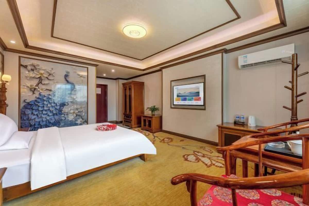Una habitación de hotel con cama, una silla de madera, una mesita pequeña, aire acondicionado de pared y un cuadro enmarcado.