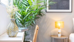 Un escritorio blanco con un florero estampado que contiene flores blancas, una planta verde y un cuadro en la pared.