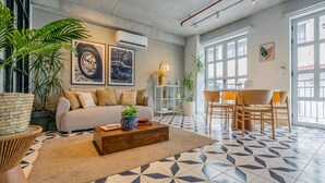 Amplia sala de estar con sofá, mesa de centro y zona de comedor. Cuenta con ventanas grandes, techo con tuberías a la vista y varias plantas.