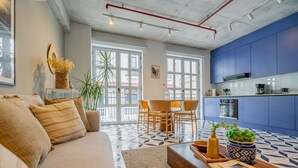 Una sala de estar con un sofá, una mesa de centro y un comedor con sillas. Hay una cocina con gabinetes azules y una ventana con vista a la calle.