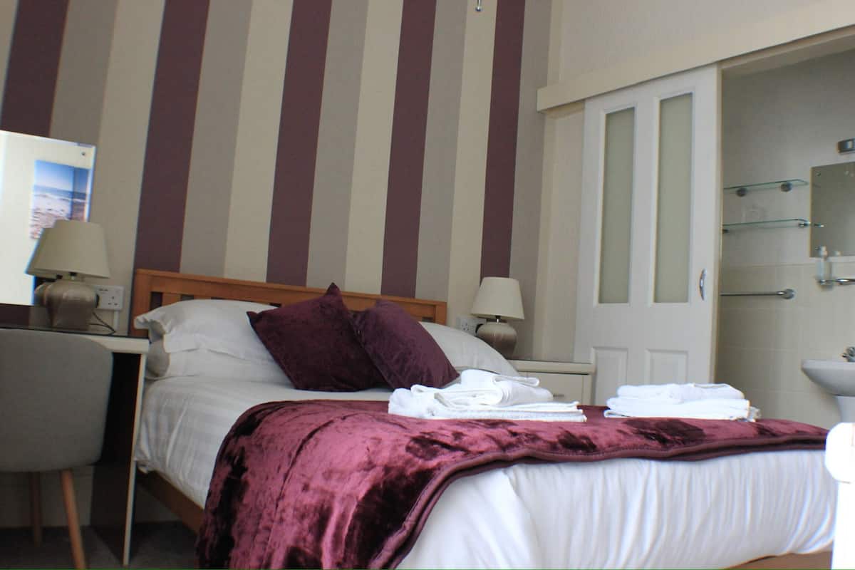 A hotel room with a bed, two pillows, a bedside table with lamps, a chair, a desk, and a bathroom with a sink and mirror.