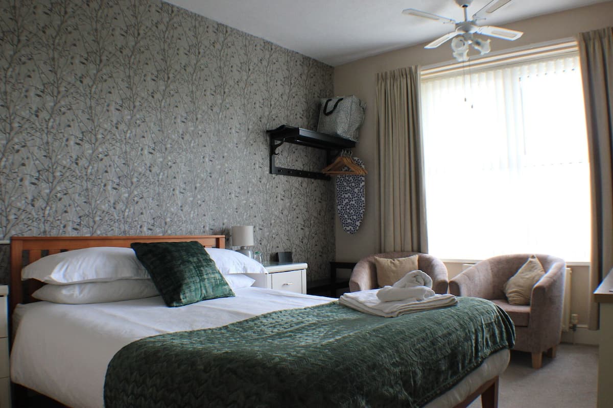 A hotel room with a bed, a chair, a nightstand, a lamp, a ceiling fan, and a window with curtains.