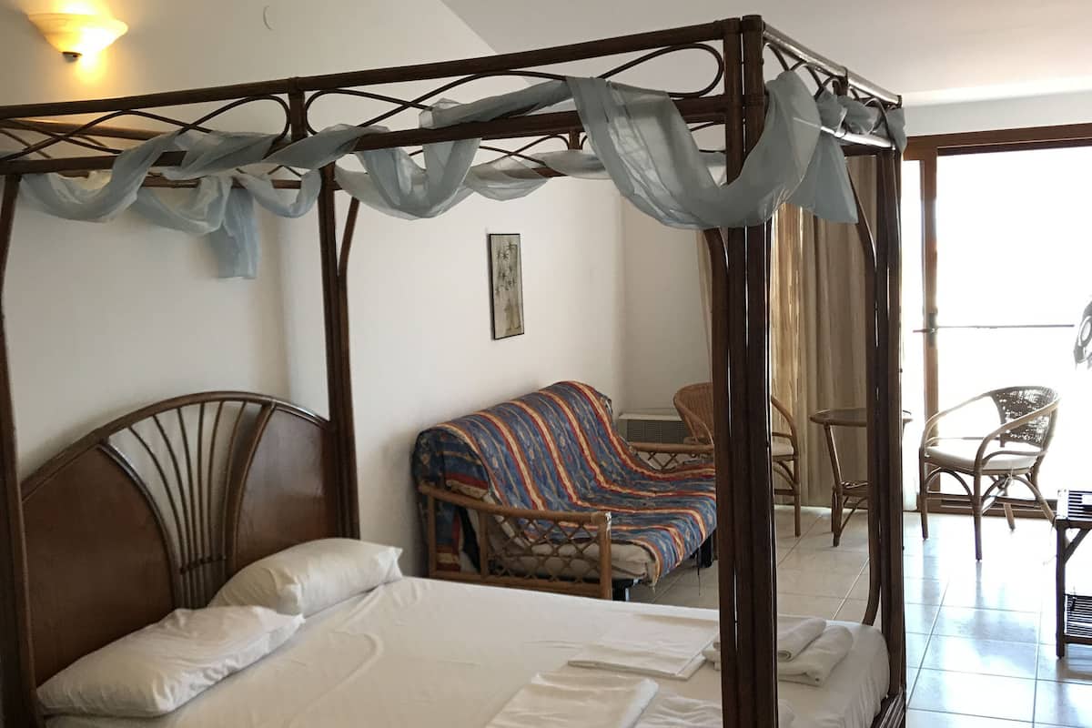 A four-poster bed with a canopy, a sofa, and a chair in a room with a tiled floor and a view through a glass door.