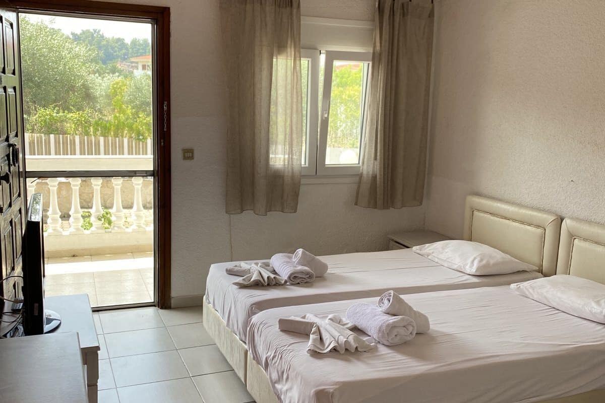 A hotel room with two beds, a desk, and a balcony with a view of greenery.