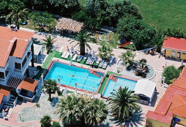 An aerial view of a resort with a central swimming pool, lounge chairs, palm trees, and a thatched-roof structure. - Villa George (Kassandra)