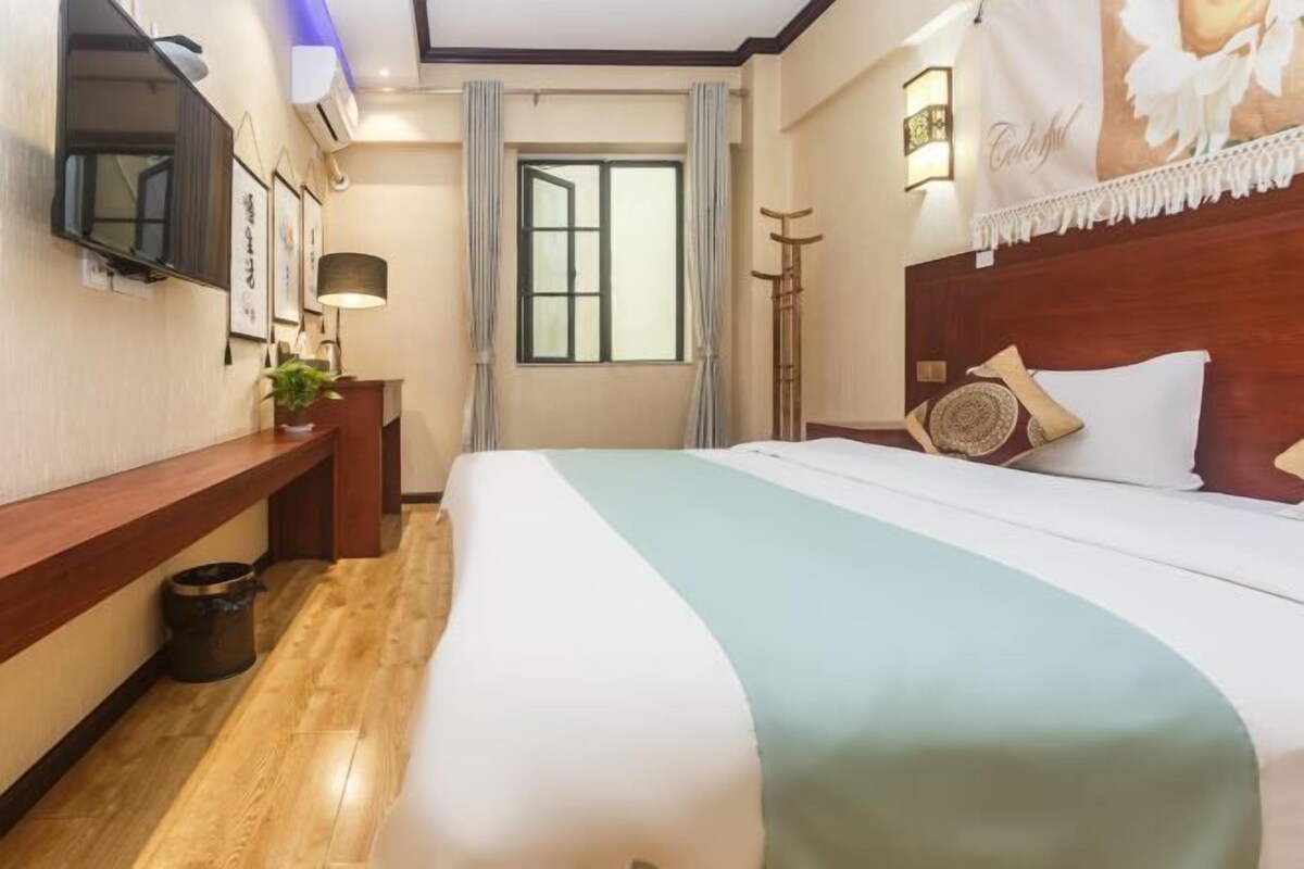 A hotel room with a bed, a desk, a lamp, a window, and a wall-mounted TV.