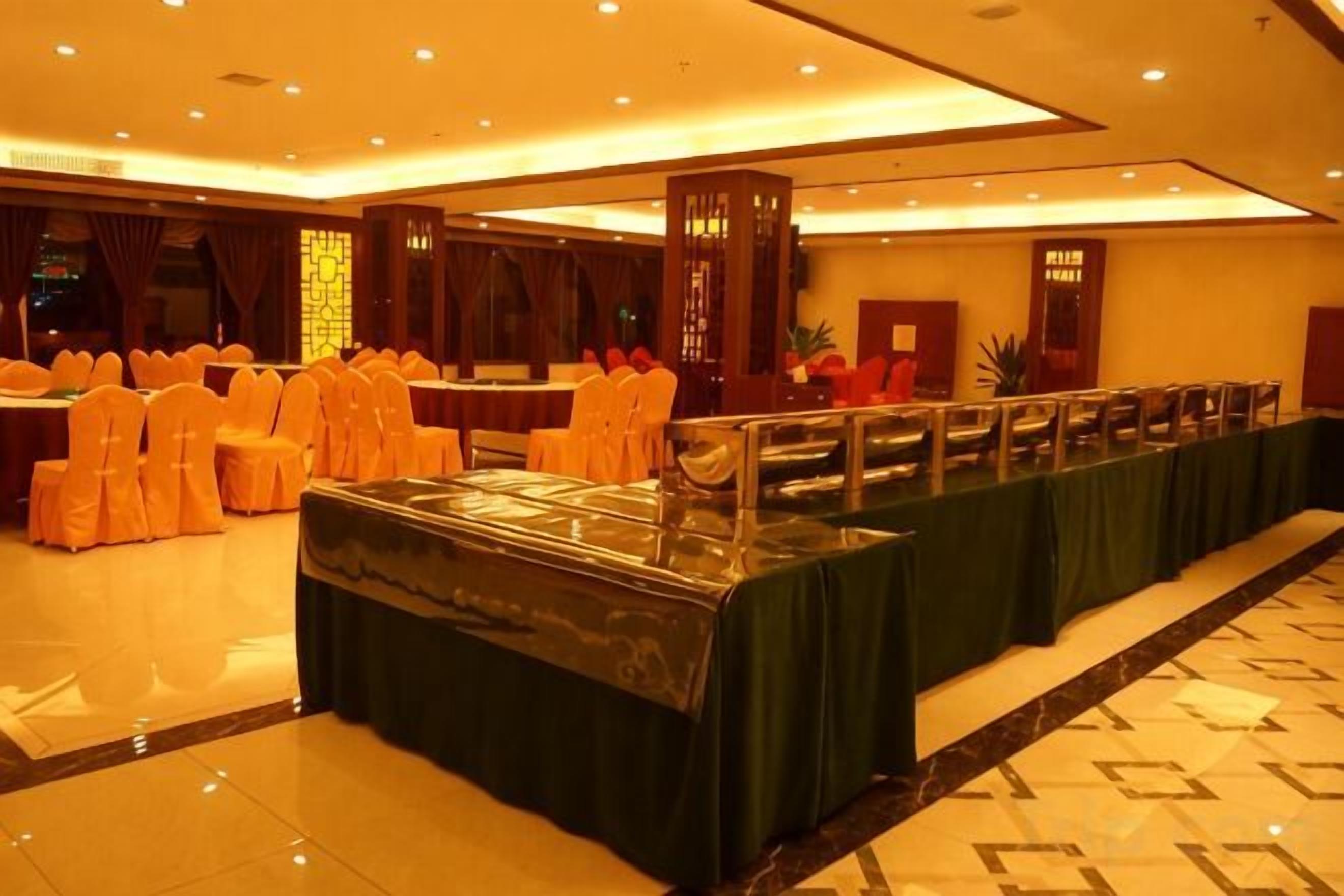 Un salón de banquetes con una larga mesa cubierta con mantel verde, filas de sillas con cintas anaranjadas y un suelo de baldosas.