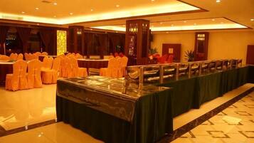 Un salón de banquetes con una larga mesa cubierta con mantel verde, filas de sillas con cintas anaranjadas y un suelo de baldosas.