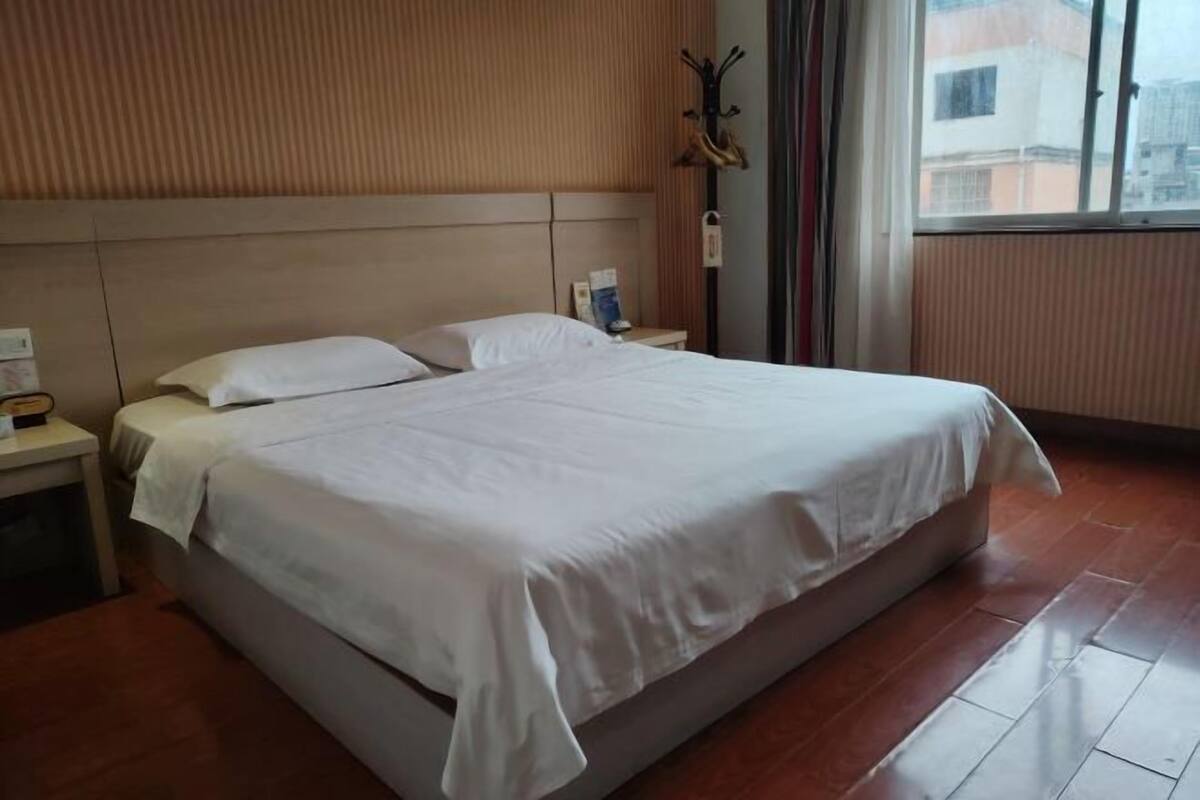 A hotel room with a large bed, wooden headboard, a nightstand with a lamp, and a window with a view of buildings.