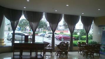 Un hall de hotel con un banco y sillas, ventanas grandes con cortinas y vista a la calle.