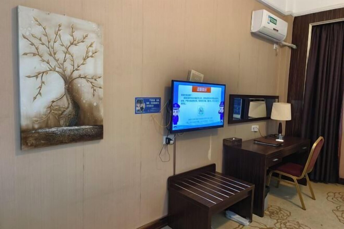 A hotel room with a flat-screen TV mounted on the wall, a desk with a chair, a small bench, and a painting of a tree.