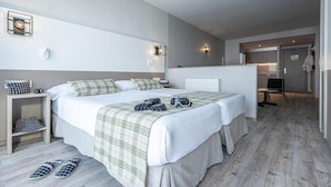 Une chambre d’hôtel moderne avec un grand lit, une table de chevet équipée d’une caméra, une paire de chaussons et une petite étagère.