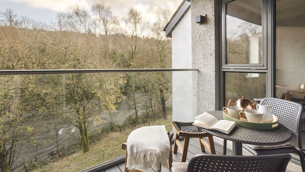 Un balcon avec une table préparée pour le thé, une chaise et une vue sur une rivière et des arbres.