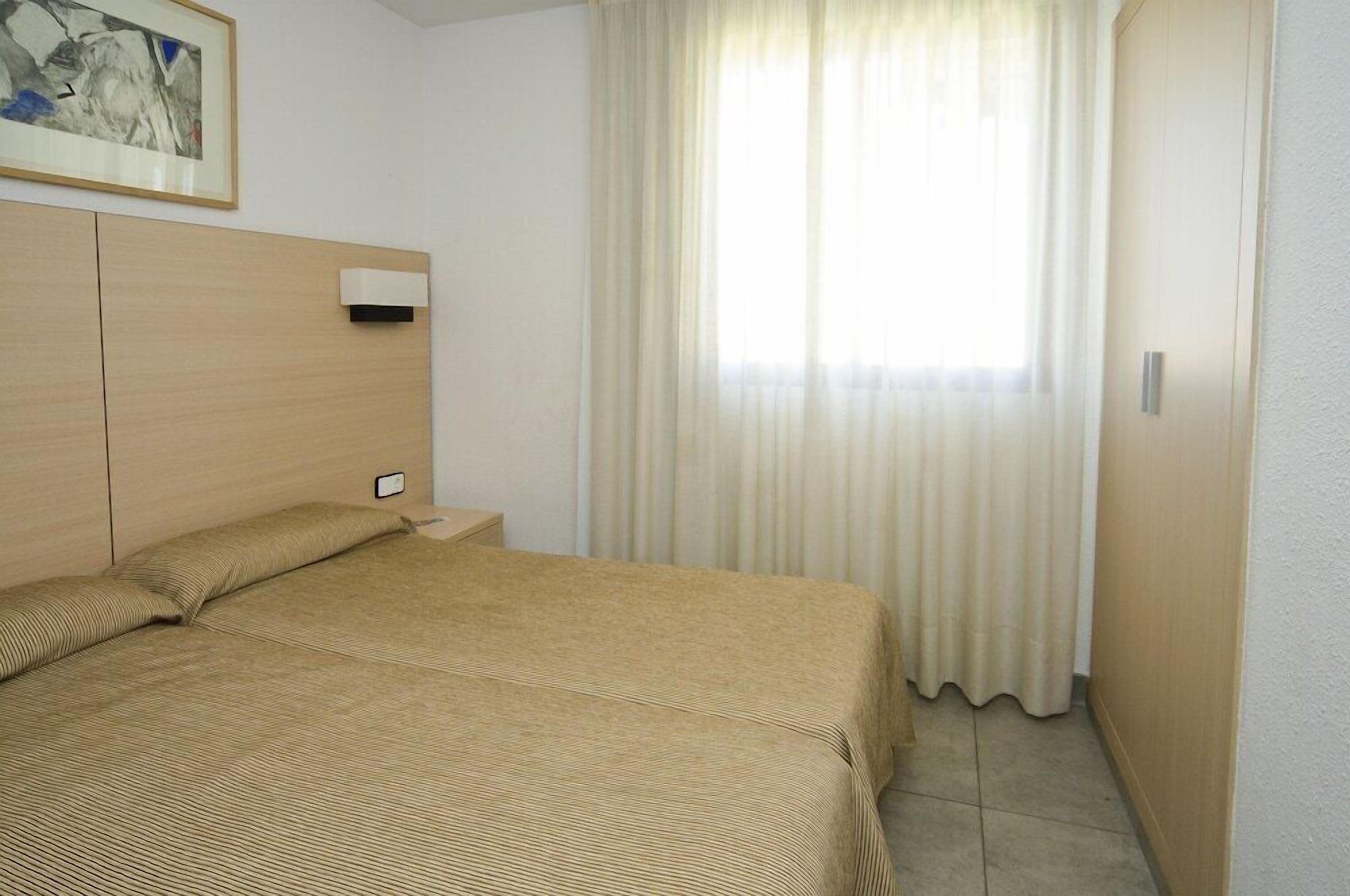 A hotel room with a bed, a nightstand, a wall-mounted air conditioning unit, and a window with curtains.