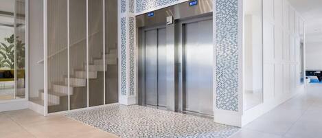 Un ascensor moderno con detalles metálicos, un felpudo estampado y una escalera con barandillas de vidrio.