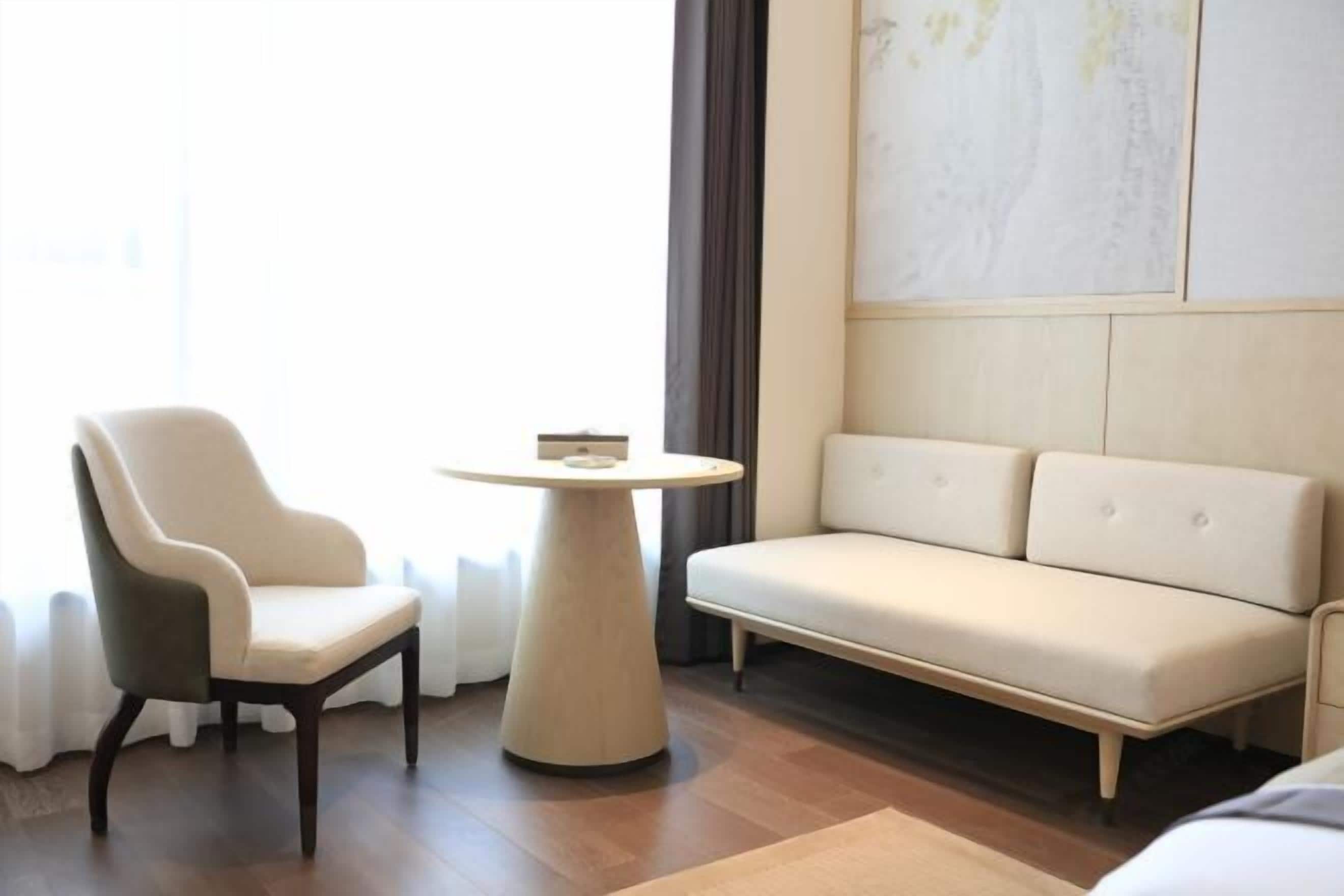 Habitació d'hotel moderna amb un sofà beix, una cadira de fusta fosca i una taula rodona petita.
