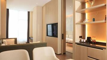 A modern hotel room with a flat-screen TV mounted on the wall, a sofa, a bed, and a small kitchenette area with a coffee maker and bottles.