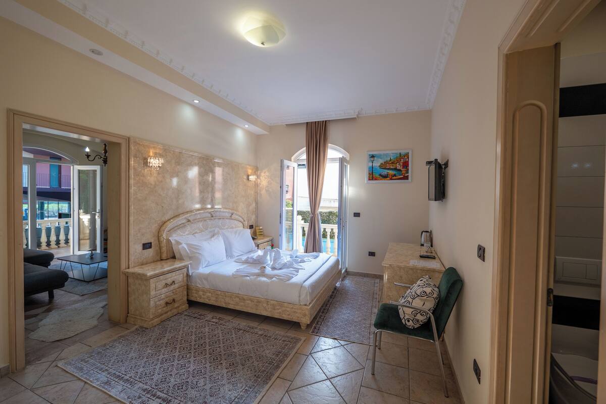 Ein Schlafzimmer mit einem großen Bett, einem Nachttisch, einem Sessel, einem Schreibtisch und Blick auf den Balkon.