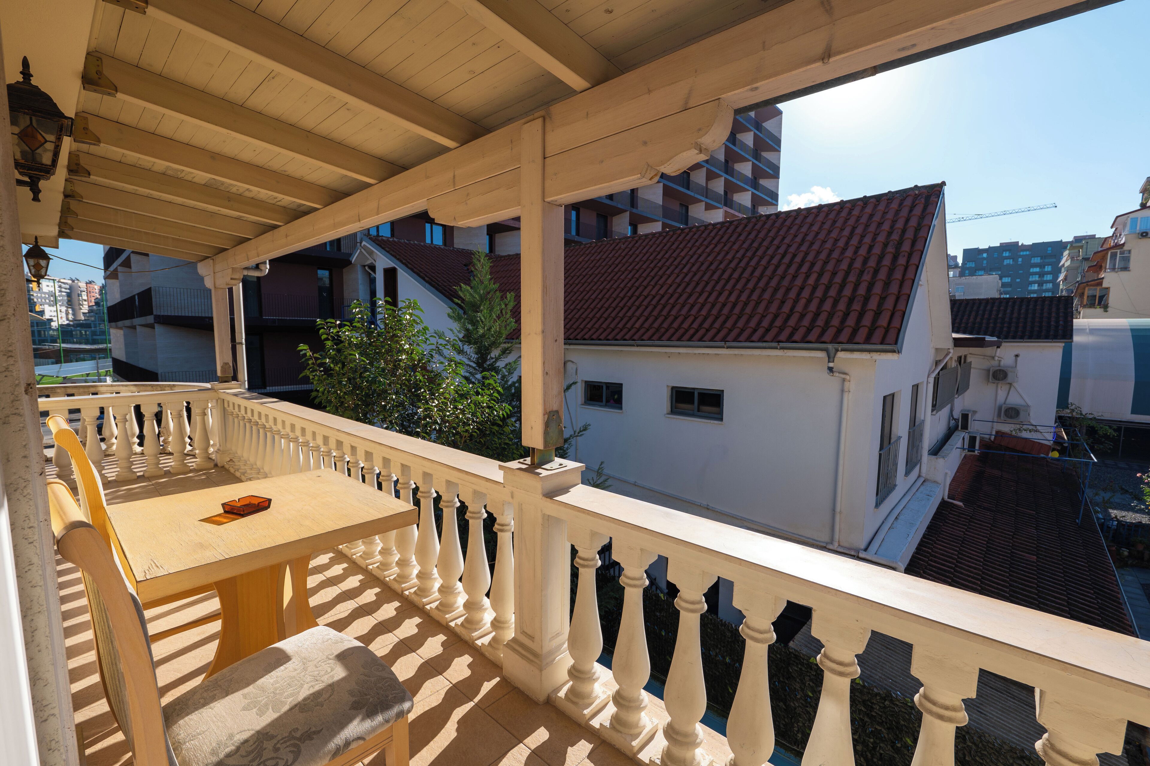 A covered balcony with a wooden table and chairs, a white balustrade, and a view of surrounding buildings.