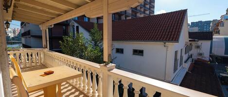 A covered balcony with a wooden table and chairs, a white balustrade, and a view of surrounding buildings.