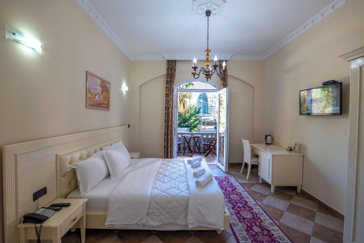 Habitación de hotel con cama, escritorio, silla, televisor y balcón con vista a edificios y vegetación.
