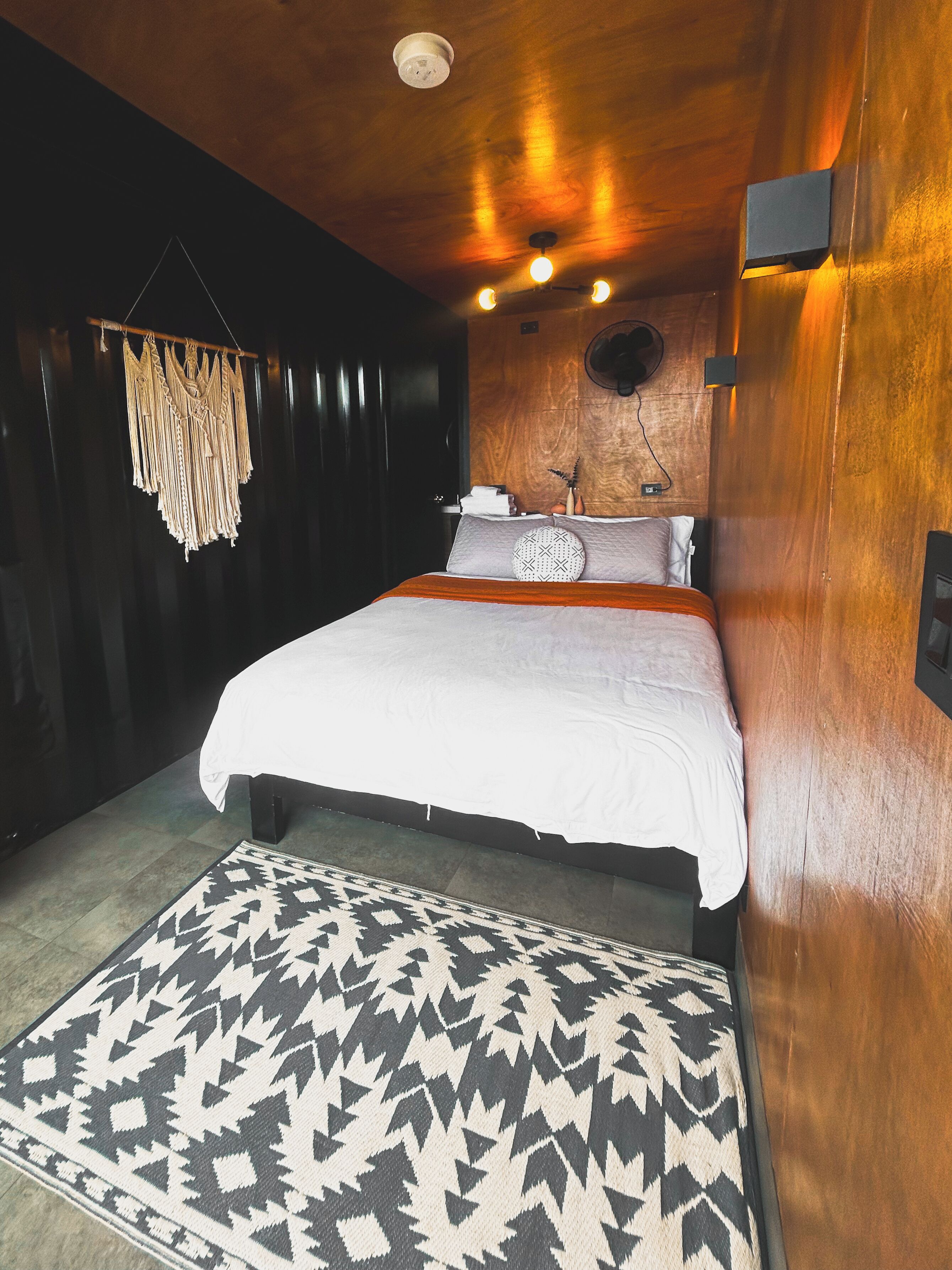 A bedroom with a bed, a patterned rug, a wall-mounted light fixture, and a ceiling fan.