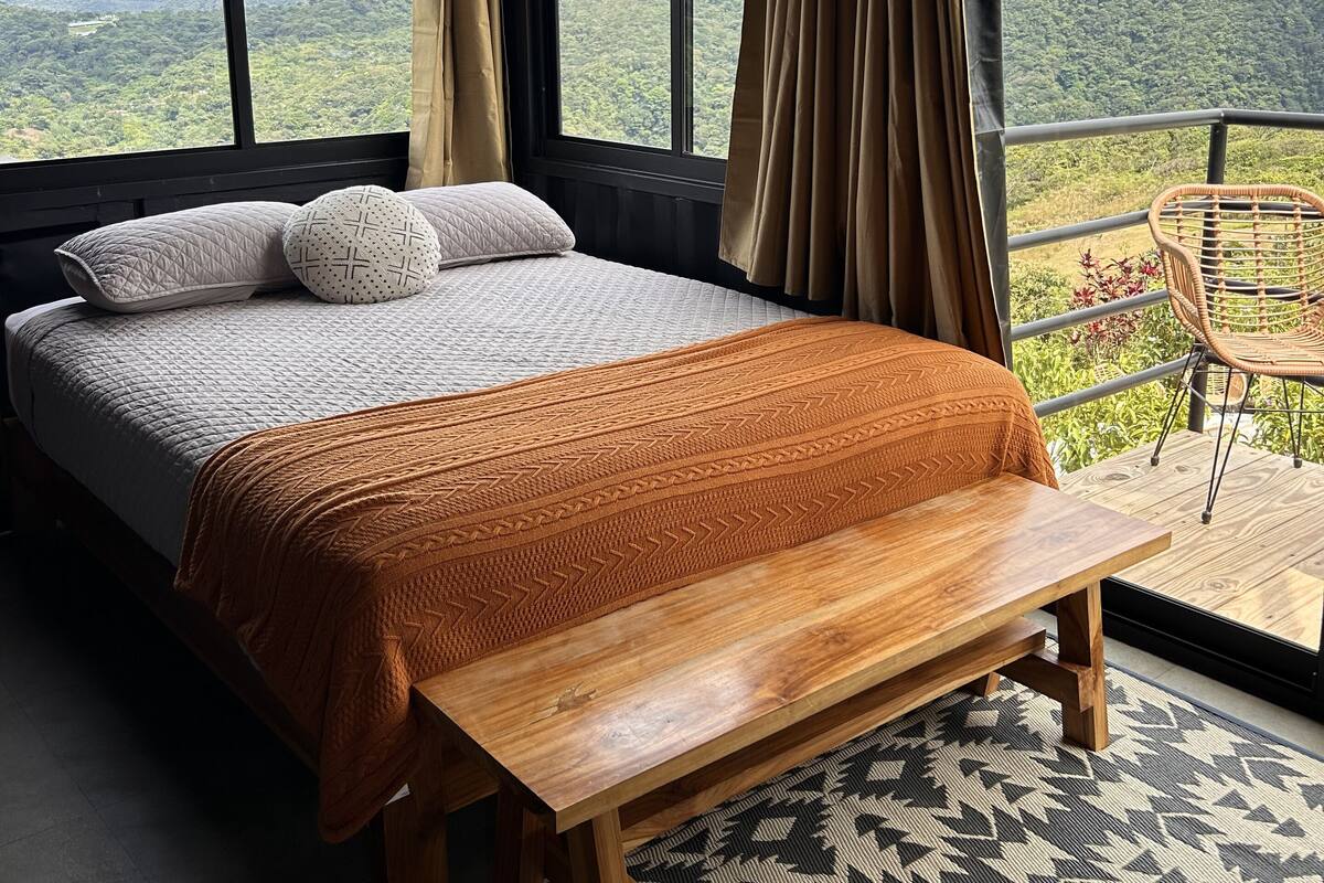 Une chambre avec un grand lit, un banc en bois et une vue sur les montagnes, visible à travers de grandes fenêtres.