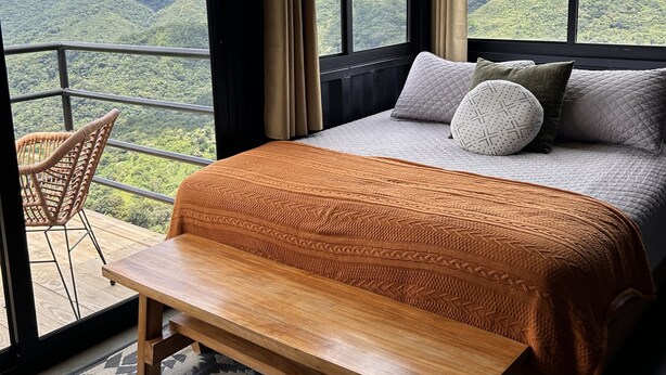 A bedroom with a large bed, a wooden bench, and a view of a mountainous landscape through large windows. - Nomada Republic Hotel El Valle  (El Valle de Anton)